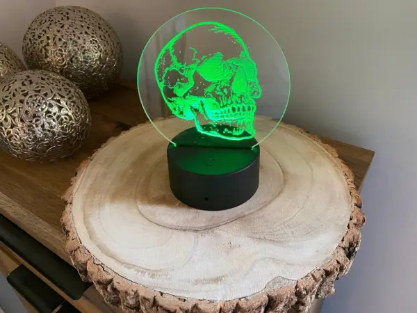 An Anatomical Skull LED Lamp - Multi-colours with remote and USB or battery powered, glows atop a round wooden slab on a wood surface, next to two decorative metal spheres with intricate cut-out designs.