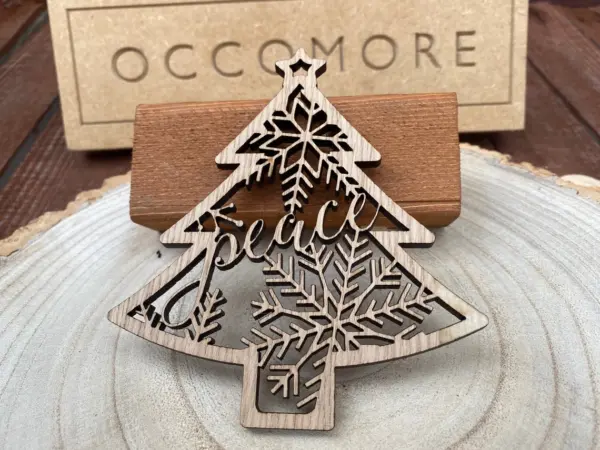 The Peace Christmas | Xmas Tree Ornament is a laser-cut wooden piece shaped like a tree with peace and snowflake designs, displayed on a round base with an OCCOMORE box in the background.