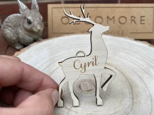 A hand holds a Personalised Christmas Reindeer Decorations Hanging Ornament (Maple or Oak) engraved with “Cyril” on a tree slice. A ceramic rabbit and a cardboard sign sit in the background, adding to its charm.