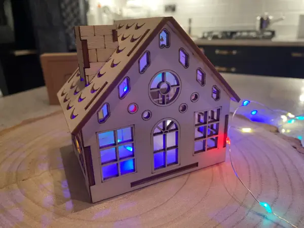 The Christmas House | Christmas Village House Kit features a small wooden Xmas house with cut-out windows that glows with colorful lights, perfect for display on a table or surface in any modern kitchen or home.