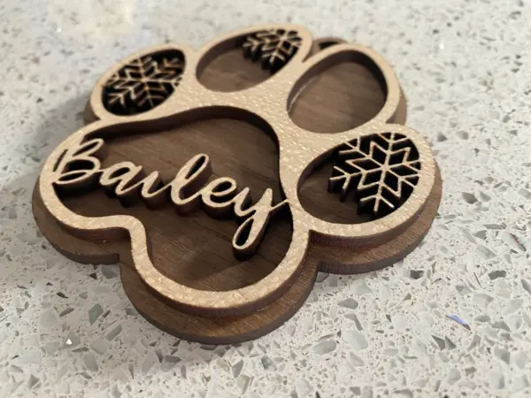 The Personalised Dog Paw Print Christmas Bauble | Tree Decoration features a custom name in cursive and three snowflakes, making it the perfect festive ornament for your tree or as a Christmas bauble. Shown here on a speckled countertop.