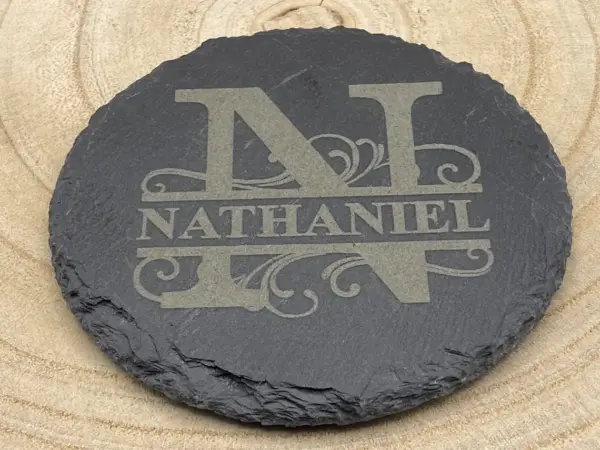 A round black Split Name Slate Coaster Monogram, personalised with a large engraved letter N and the name NATHANIEL, displayed on a light wood surface with circular grain patterns.