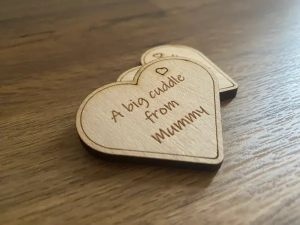 A wooden heart-shaped A Big Cuddle from Mummy Token—customizable for Daddy, Auntie, Nanny, etc.—rests on a wooden surface. Its a charming First Day At School or Starting Nursery Keepsake; another token is partially visible in the background.