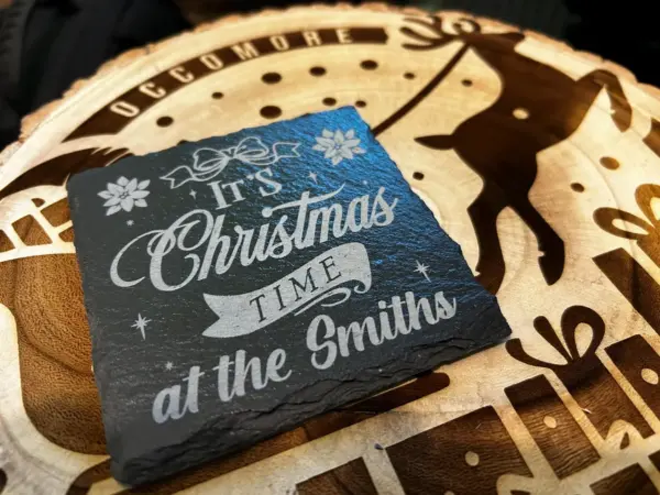 A matt finish personalised slate coaster, available in circle or square, reads Its Christmas Time at [Names], and rests on a wooden surface with an engraved reindeer and festive patterns.