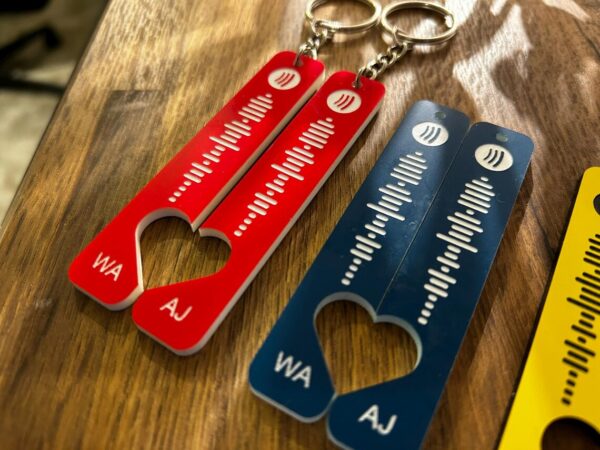 Two Wedding Song Keyrings—one red, one blue—each featuring your chosen Spotify Code engraving, cut-out heart design, and initials WA and AJ at the bottom. Two tags with chains are shown on a wooden surface.